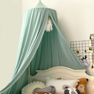 Grüne Babywiege hängt über einem weißen Bett mit weißen, beigefarbenen und grauen Kissen vor einer weißen Wand