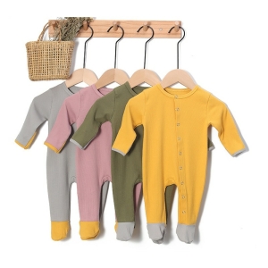 Gerippter Babystrampler aus gelber, grüner, rosa und grauer Baumwolle, aufgehängt mit einem Weidenkorb an Kleiderbügeln auf einer beigen Wandgarderobe