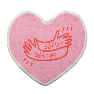 Herzförmiger Teppich für das Mädchenzimmer in rosa Farbe. Auf der Oberseite ein Druck von zwei Händen mit der Aufschrift self love self care in roter Farbe.