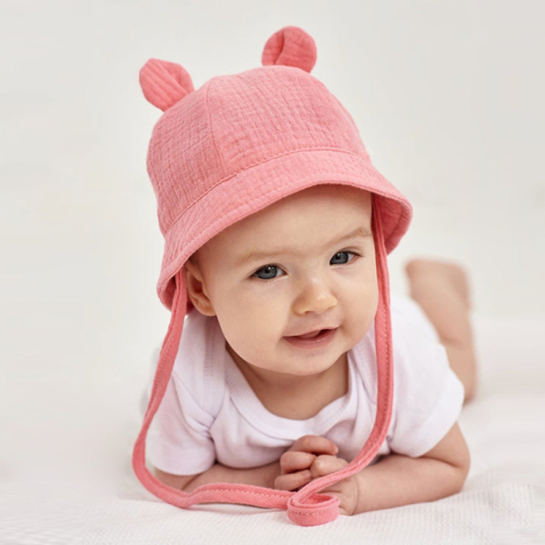 Baby-Mädchen in Bauchlage auf einem weißen Wabenstoff mit einem weißen Body und einem rosa Chiffonhut mit Ohren und Kordel.