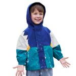 Ein kleiner kaukasischer Junge in einem dünnen Patchwork-Parka mit Kapuze in Grün, Blau, Weiß und Gelb auf einfarbigem Hintergrund.