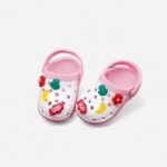 Mini-Clogs im Crocs-Stil in Rosa und Weiß mit bunten Charms oben drauf, vor neutralem Hintergrund.
