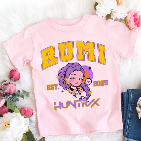 K-pop Demon Hunters Mädchen T-Shirt rosa von Charakter Rumi mit ihrem Vornamen in gelben Großbuchstaben geschrieben sowie Huntrix. Dazwischen das Gesicht in Cartoon-Version von Rumi. Das T-Shirt liegt neben Blumen.